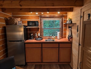 A compact kitchen with wooden walls, a refrigerator, a microwave, a coffee maker, and a window with blinds.