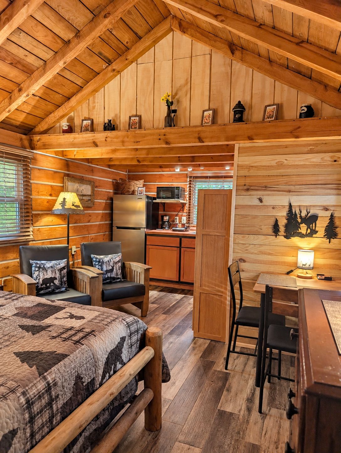 Interior acogedor de una cabaña con cama, dos sillones, una mesa de comedor, zona de cocina y techo de madera.