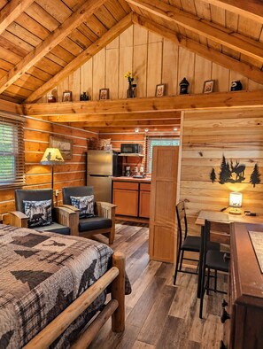 A cozy cabin interior with a bed, two chairs, a dining table, a kitchen area, and a wooden ceiling.