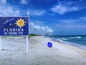 Eine Strandansicht mit einem Schild, auf dem 'Welcome to Florida the Sunshine State' steht, und einer Möwe, die darauf sitzt.