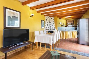 Ein Wohnzimmer mit Fernseher, einem Esstisch mit Stühlen, einer Küche im Hintergrund und einer Decke mit Holzbalken.