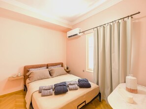 A bedroom with a bed, pillows, a bedside table, an air conditioning unit, a window with curtains, and a small table with a vase. - Ivelia Athens - Boutique Residence with Private Spa (Athina)