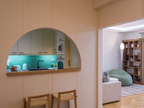 Une cuisine moderne avec un grand miroir en arc de cercle, des chaises en bois et une vue sur le salon, où l’on aperçoit un canapé et une bibliothèque.
