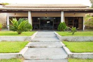 A building with a tiled roof, a central entrance with steps, and a small outdoor seating area with chairs and tables. - Casa 2 suites-piscina priv-Picos161m (Icapuí)