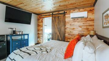 Ein Schlafzimmer mit Holzbalkendecke, einem Bett mit weißen und orangefarbenen Kissen, einer blauen Kommode und einem an der Wand befestigten Flachbildfernseher.