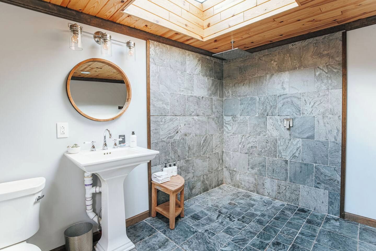 A bathroom with a white pedestal sink, a round mirror, and a shower area with stone tiles.