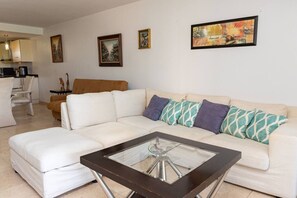 Ein Wohnzimmer mit einem weißen Sofa, einem Glastisch und einer Küche im Hintergrund.