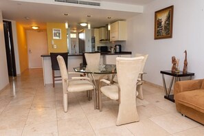 A dining area with a glass table and cushioned chairs, a kitchen in the background, and a painting on the wall.