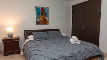 Um quarto com cama, mesa de cabeceira, abajur, quadro e guarda-roupa.