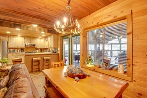 A wooden interior with a dining table, bar stools, a chandelier, and a view through a window.