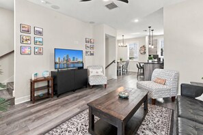 A modern living room with a flat-screen TV, a sofa, a coffee table, and a dining area with a kitchen in the background. - River District Residence | Walk to Armature Works | 3BR Townhome with Garage & Private Yard (Tampa)