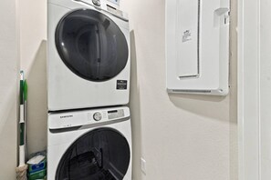 A wall-mounted washing machine and dryer, with a green-handled mop and detergent boxes nearby. - River District Residence | Walk to Armature Works | 3BR Townhome with Garage & Private Yard (Tampa)