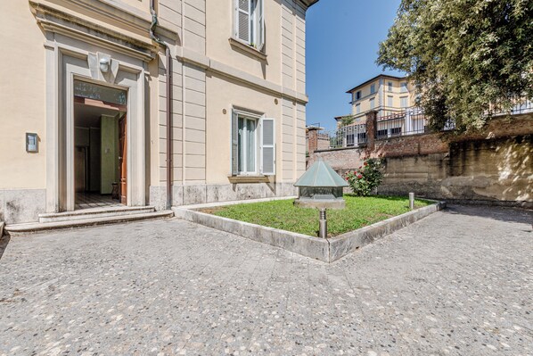 A building with a paved courtyard, a small garden area with a metal lamp post, and a door leading to an interior space.