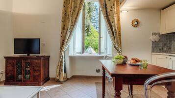 Ein Zimmer mit Fernseher auf einer Holzkommode, einem Esstisch mit Obstschale und Glas sowie einem Fenster mit Vorhängen.