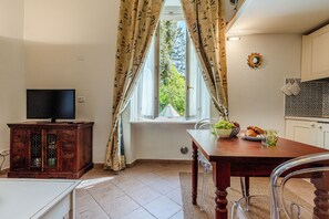 A room with a television on a wooden cabinet, a dining table with a bowl of fruit and a glass, and a window with curtains.