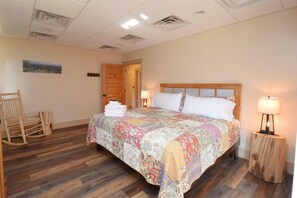 Ein Hotelzimmer mit Holzboden, einem Bett mit blumigem Überwurf, zwei Nachttischlampen, einem Holzschaukelstuhl und einer Tür zu einem anderen Raum.