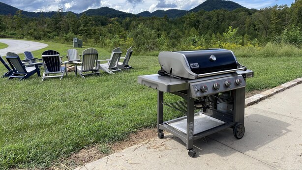 Een draagbare grill met een metalen kar, vier Adirondack-stoelen, en een mooi uitzicht op bergen en bomen.