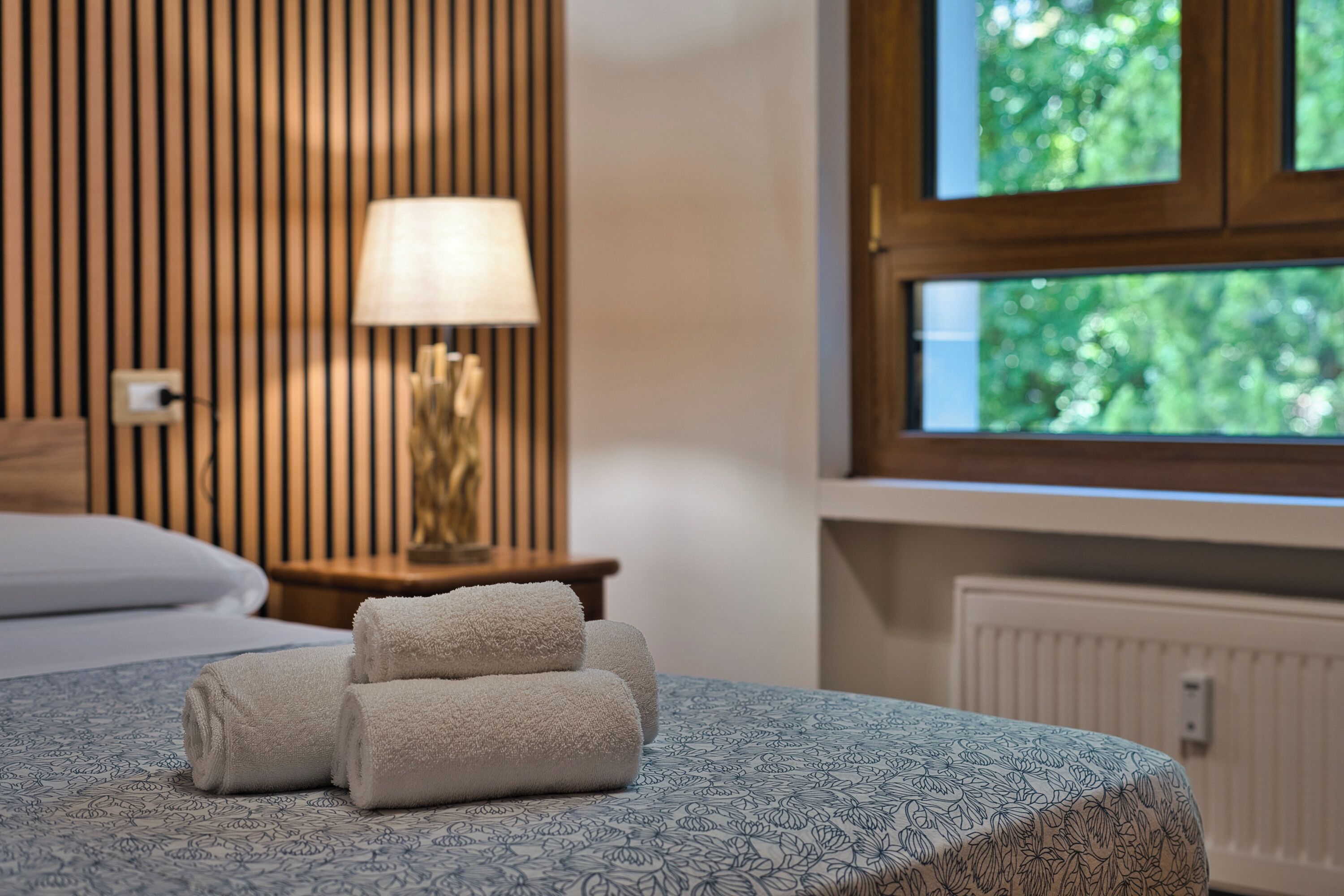 Ein ordentlich bezogenes Bett mit aufgerollten Handtüchern, einer Nachttischlampe und einem Fenster mit Blick auf Grünflächen.