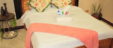 A neatly made bed with a pink towel, two floral pillows, and a small basket with toiletries.