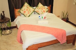 Uma cama bem arrumada com uma toalha rosa, dois travesseiros florais e uma cesta pequena com artigos de higiene pessoal.
