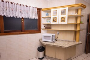 A compact kitchen with a microwave, a trash bin, and a window with a lace curtain.