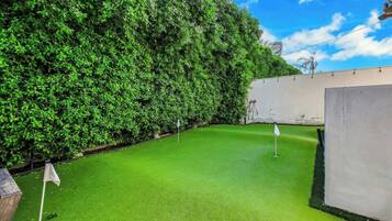 A green artificial turf area with a flagstick, surrounded by dense foliage and a white wall.