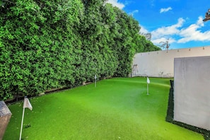 A green artificial turf area with a flagstick, surrounded by dense foliage and a white wall.