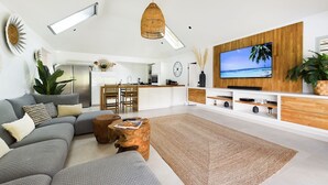 Uma sala de estar moderna com uma grande televisão de tela plana, um sofá seccional cinza, uma mesa de centro e uma área de cozinha ao fundo.
