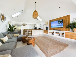 Ein modernes Wohnzimmer mit einem großen Flachbildfernseher, einem grauen Ecksofa, einem Couchtisch und einer Küche im Hintergrund.