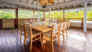 Um deck de madeira com uma mesa posta para refeições, rodeado por cadeiras de vime, sob uma área coberta com vista para o verde.