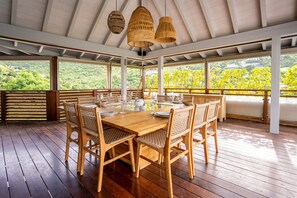 Eine Holzterrasse mit Esstisch, umgeben von Korbstühlen, unter einem Überdachung mit Blick auf Grünflächen.