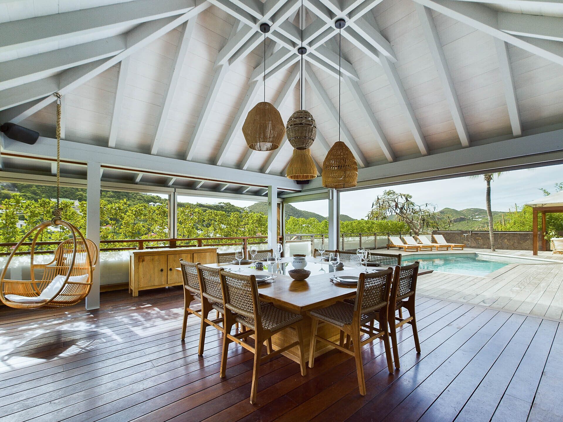 An outdoor dining area with a wooden table and chairs, surrounded by a wooden deck and overlooking a pool and lush greenery.