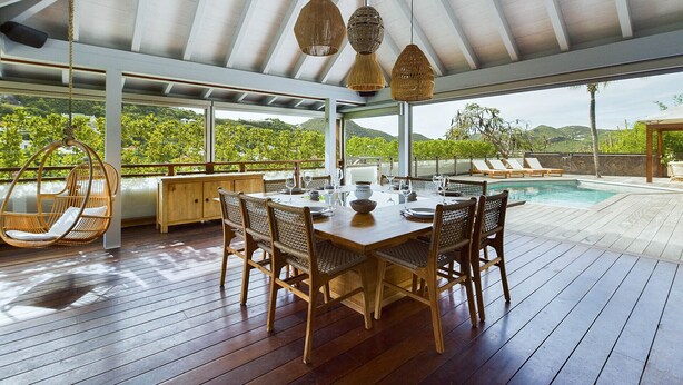 Uma área de refeições ao ar livre com mesa e cadeiras de madeira, rodeada por um deck de madeira e com vista para uma piscina e vegetação exuberante.