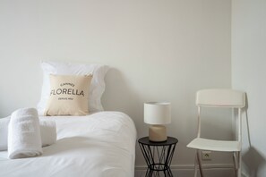 Ein ordentlich bezogenes Bett mit einem Kissen, auf dem 'CANNES FLORELLA DEPUIS 1952' steht, eine Nachttischlampe und ein weißer Stuhl.