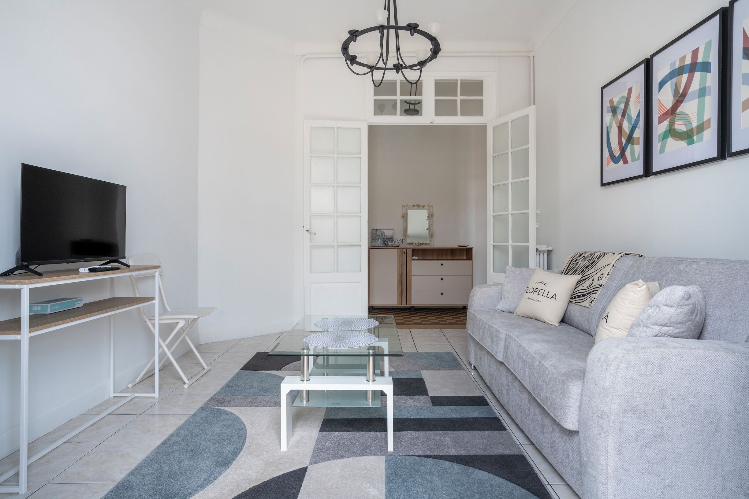 A modern living room with a flat-screen TV, a glass coffee table, a grey sofa with decorative pillows, and a chandelier.