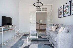 Ein modernes Wohnzimmer mit einem Flachbildfernseher, einem Glastisch, einem grauen Sofa mit Zierkissen und einer Chandelier.