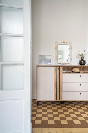 Ein Zimmer mit Schachbrettboden, einer weißen Tür, einem Schminktisch mit Spiegel, einem Bilderrahmen und einer Vase mit Blumen.