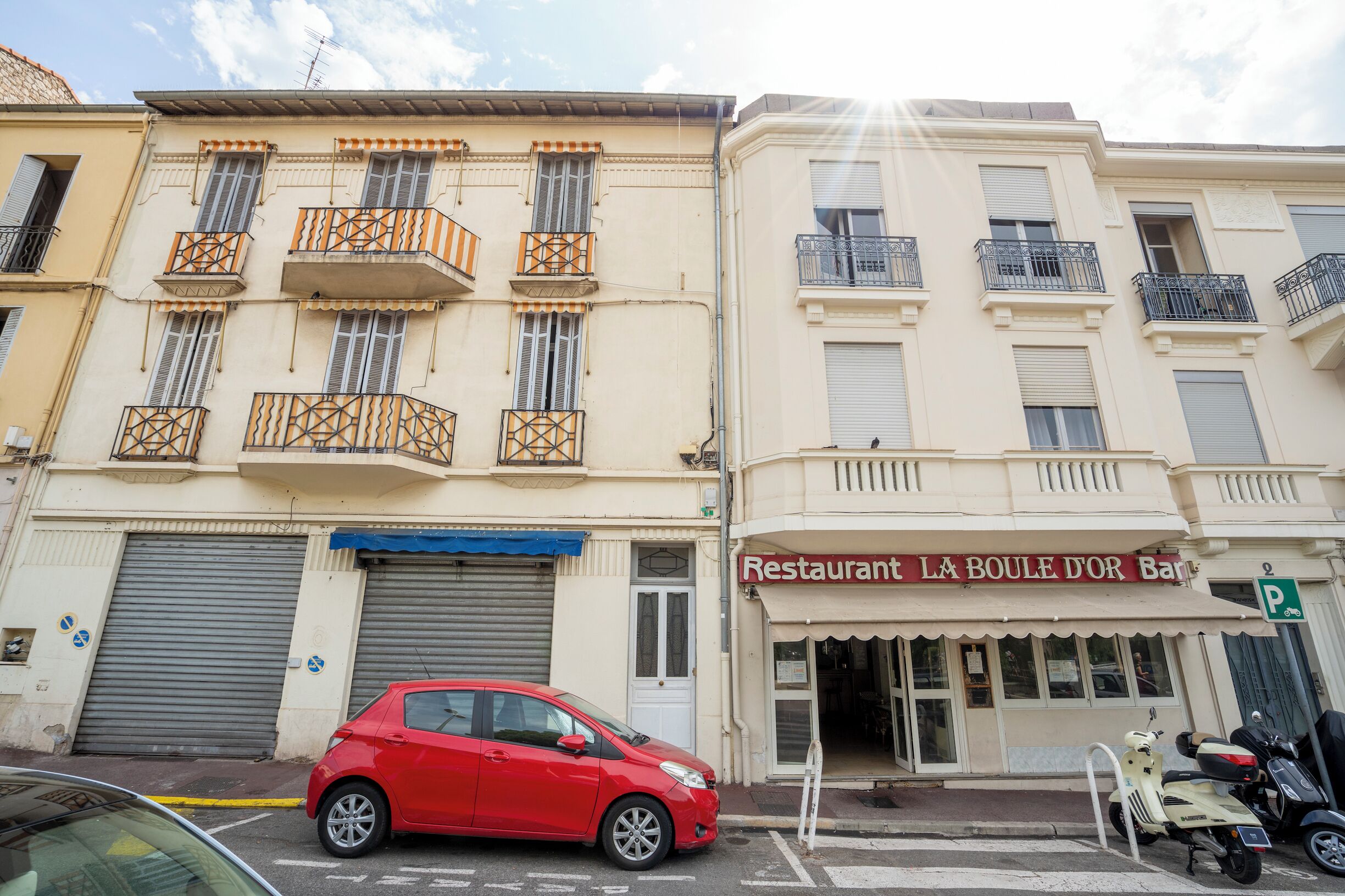 Un'auto rossa parcheggiata davanti a un edificio con un ristorante chiamato 'LA BOULE D'OR BAR' e un segnale stradale con la lettera 'P'.