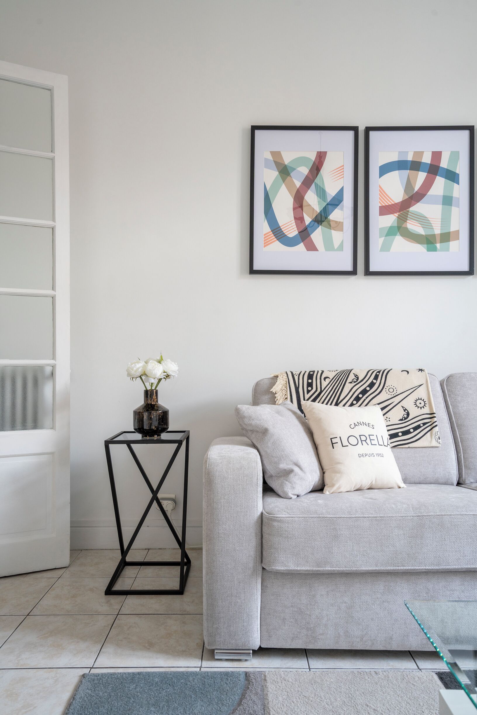 A modern living room with a grey sofa, two abstract paintings, a vase with white flowers, and a black metal side table.