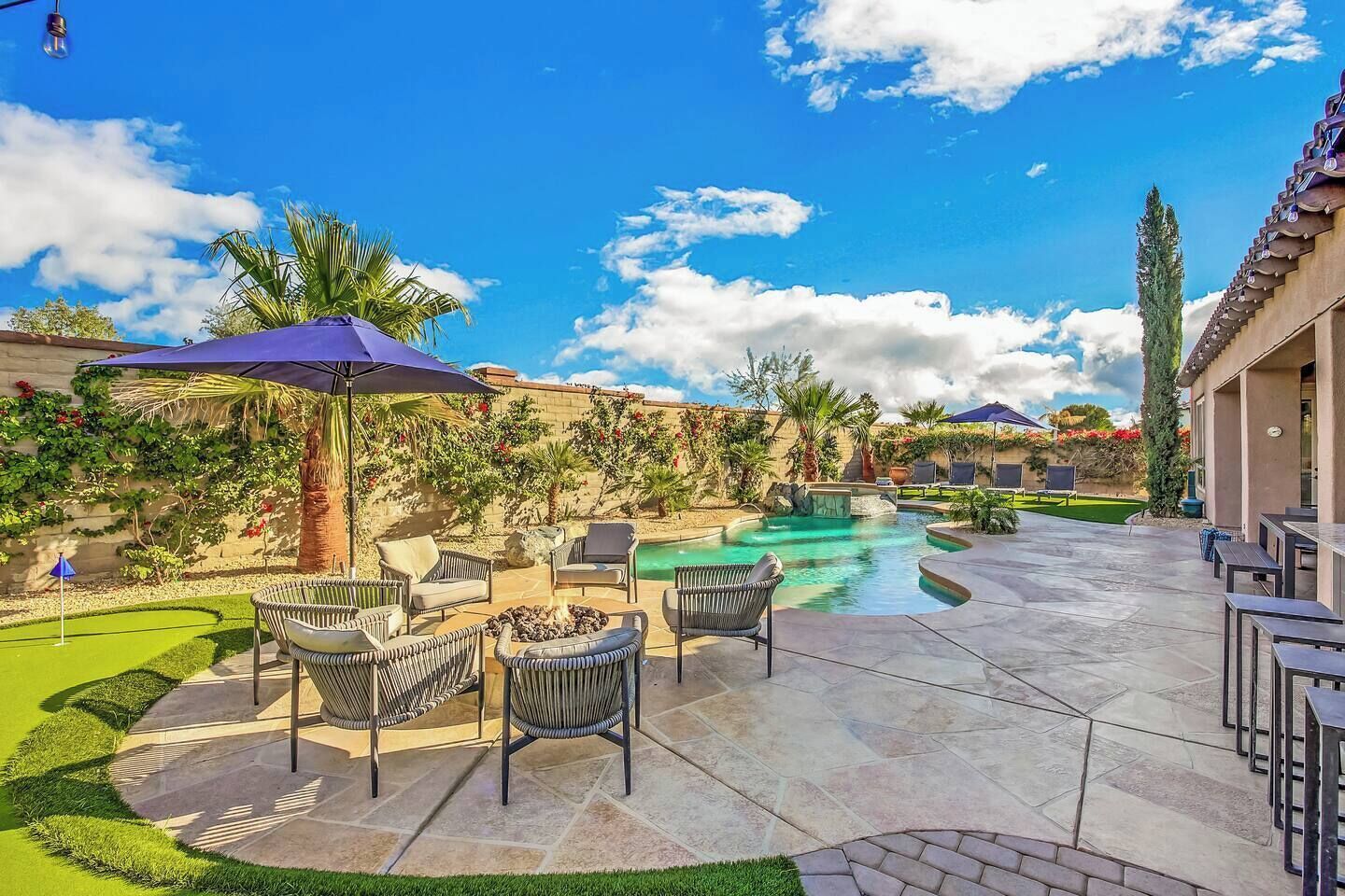 A pool area with lounge chairs and umbrellas, surrounded by landscaped gardens and a clear sky.