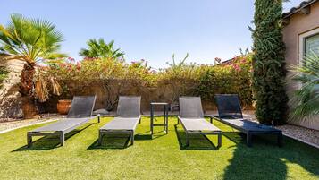 Une terrasse avec cinq chaises longues disposées en rangée, un palmier et diverses espèces d’arbustes à fleurs.
