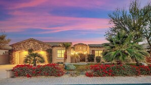 A two-story house with a stone facade, a front entrance with a door, and a well-maintained garden with red flowers and palm trees.