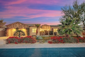 A two-story house with a stone facade, a front entrance with a door, and a well-maintained garden with red flowers and palm trees.
