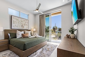 Ein modernes Schlafzimmer mit einem großen Bett, einem Deckenventilator, einem Flachbildfernseher und einem Balkon mit Blick auf Palmen und Häuser.