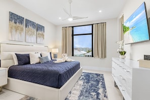 A modern bedroom with a large bed, a nightstand, a dresser, a TV, and a window with a view of buildings. - Ultimate Fort Myers Beach Getaway | 2 Adjacent Luxury Homes | 12BR/12.5BA (Fort Myers Beach)