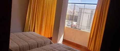 Habitación con dos camas, cortinas amarillas y vista a un paisaje urbano a través de una ventana.