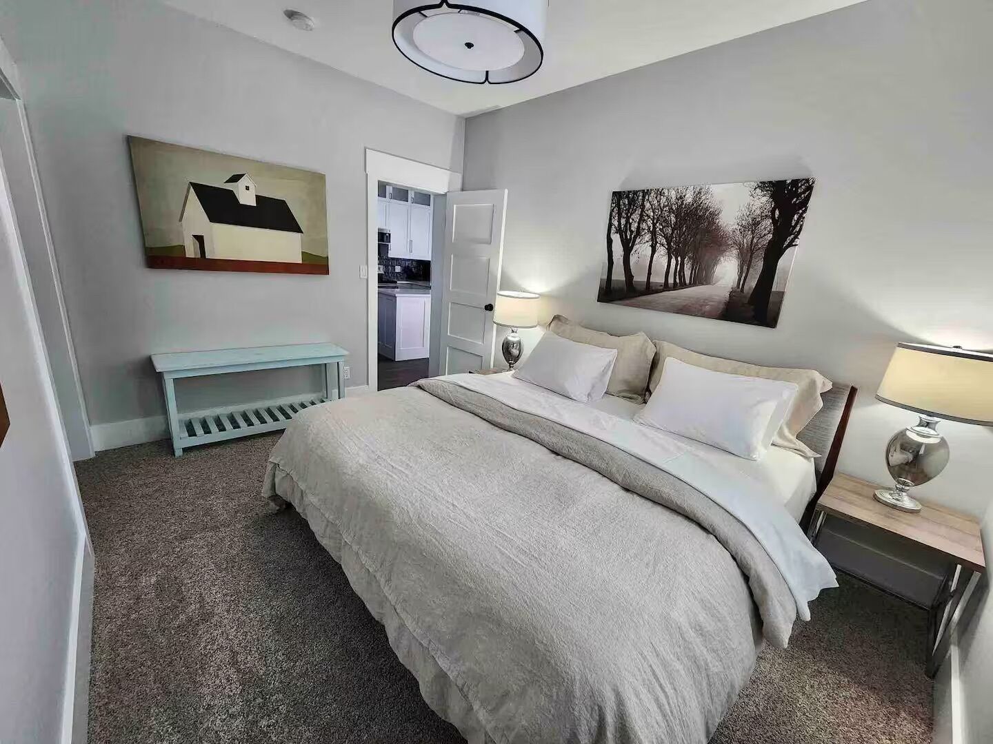A bedroom with a bed, bedside tables, a bench, a door leading to a kitchen, and a painting on the wall.