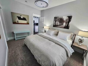 A bedroom with a bed, bedside tables, a bench, a door leading to a kitchen, and a painting on the wall. - Ski-In/Ski-Out / Private Hot Tub & Mountain Views (Kimberley)