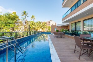 A swimming pool with a glass balustrade, surrounded by outdoor seating and a view of a modern building with large windows. - Apê Completo em Porto de Galinhas-SH164D (Ipojuca)