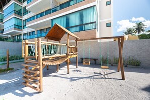 A wooden play structure with a slide and swings, located in a sandy area with a building in the background. - Apê Completo em Porto de Galinhas-SH164D (Ipojuca)
